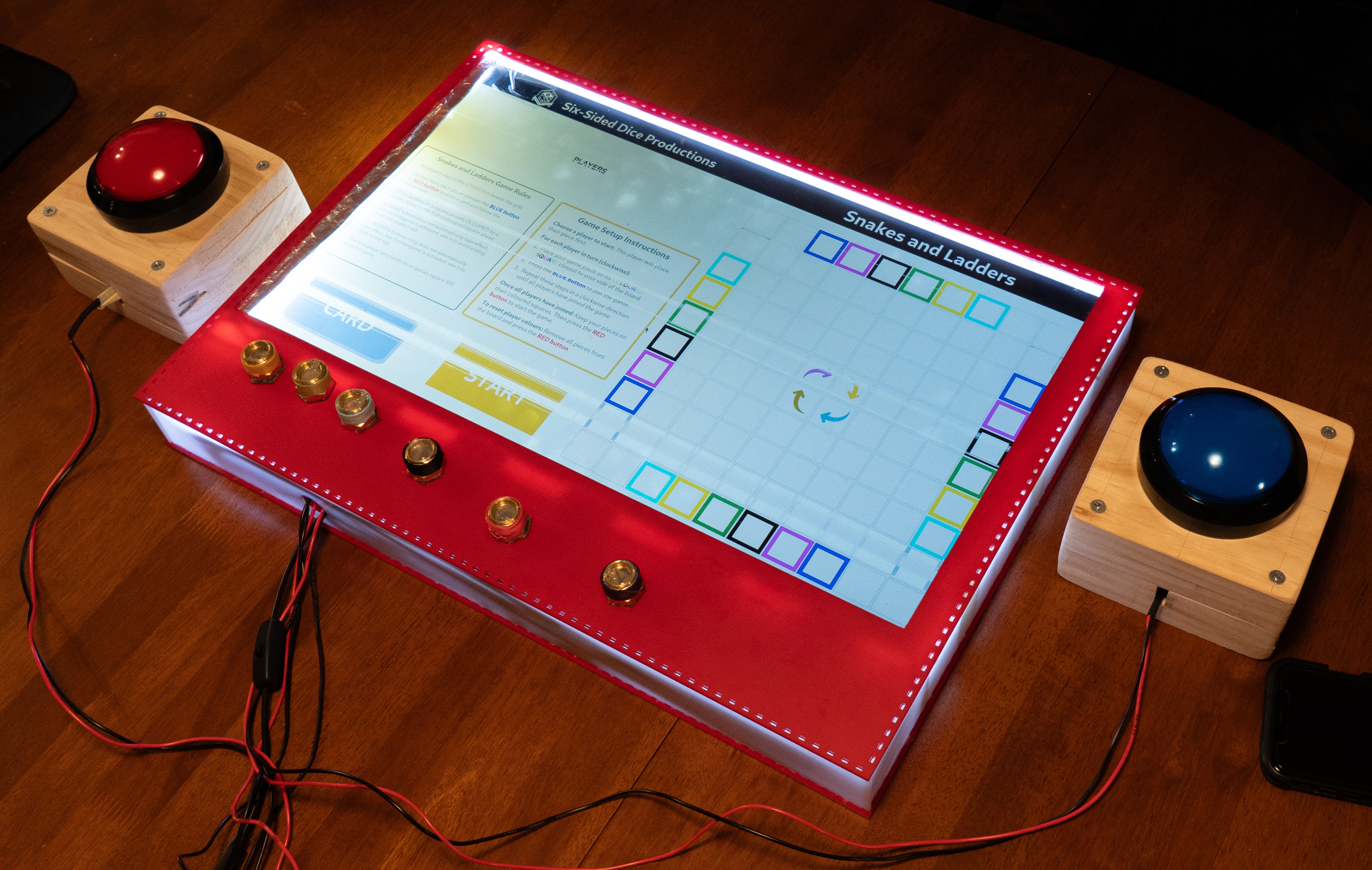 Device with screen showing a digital version of snakes and ladders. Connected via wires to a big tactile red and blue button. Six game pieces sitting on the device.